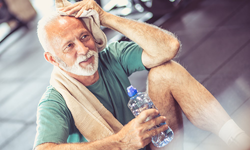 Older man sitting on gym floor with a bottle of water and towel after exercising