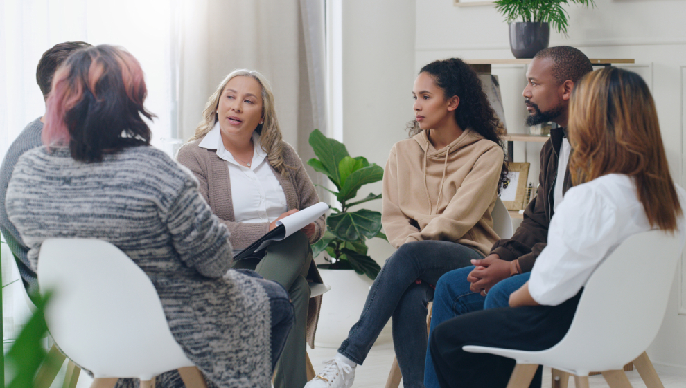 Group of people sat in a circle, including a woman with a clipboard