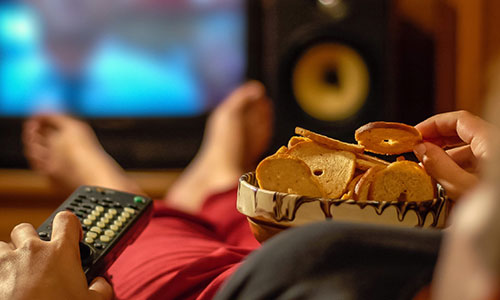 Person reclining on couch watching TV and eating from a bowl of crisps.